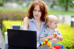 Young mother working oh her laptop and holding her sad toddler son