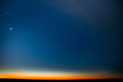 Night Starry Sky With Glowing Stars Above Countryside Field Landscape In Early Spring. Bright Glow Of Planet Venus In Sky Among The Stars. Sky In Warm Lights Of Evening Sunset Dawn Or Morning Sunrise.
