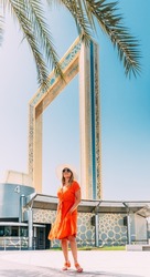 Vertical High quality photo. Young Caucasian Woman In Red Dress And In Summer Hat Standing Near Dubai Frame And Posing For Camera. View Of Dubai Frame In Zabeel Park On Sunny Blue Sky Under Palm