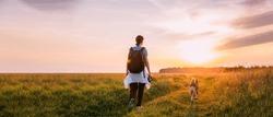 Scenic View Bright Sunbeams. Young Woman Backpacker Walking With Dog In Summer Meadow Grass During Sunset Sunrise Time. Healthy Lifestyle Walks In Evening Summer Sunlight. Panoramic View.