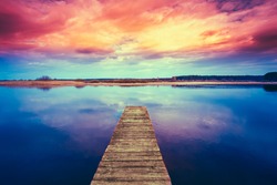 Colorful sunset dramatic sky over wooden boards pier on Calm Water Of Lake, River. Nature Background.