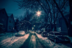 Dark city street with trees, snow and parked cars at night in the winter.