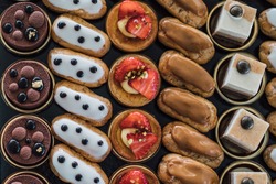 Variety of delicious confectionery products on black tray. A mix of chocolate mousse, eclair cakes and fruit tarts for a candy bar, or a pastry shop showcase presentation. Flat lay, top view dessert.