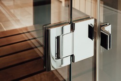 Close metal silver hinge is mounted on the glass construction of the door to the sauna, bathroom or shower cubicle against the background of the wooden floor
