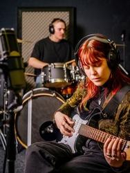 Young redhead woman playing electric guitar and a male drummer in a recording studio.