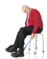 Full length view of senior adult looking at the viewer as he puts his loafers on with a long-handled shoe horn.  On a white background.