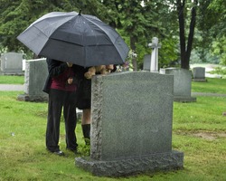Two young people under an umbrella bringing a teddy bear and flowers to a grave site in the rain.