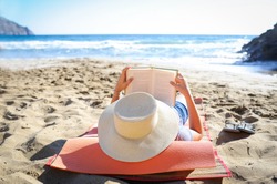 Caucasian woman is reading a book on a sandy beach wearing a straw hat