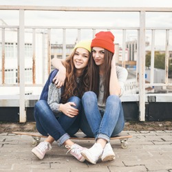 Two young girl friends sitting together on long-board and having fun. Outdoors, lifestyle.