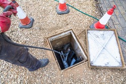 Man unblocking domestic sewage drain through open inspection chamber, drain cleaning company, UK