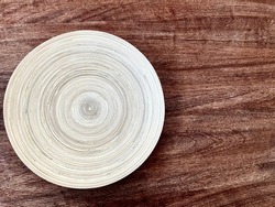 Close-up of a white wooden plate on the surface of a dark wooden tabletop. A wooden plate with a texture of concentric circles on a dark brown wooden table.