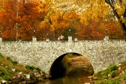 Couple stand on stone bridge and are surrounded by the blazing orange and yellow of an Autumn morning.