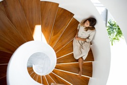 Woman in linen natural dress is sitting on the wooden modern stairs inside of the house