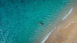 Aerial view of beautiful happy woman in swimsuit laying in the shallow sea water, enjoying sandy beach and soft turquoise ocean wave. Tropical sea in summer season on Egremni beach on Lefkada island.