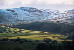 Sunrise lights the pattern of green patchwork fields and ancient field systems below a snow-capped hill near Malham, Yorkshire, England