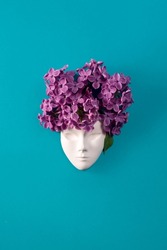 Plaster face, figurine decorated with lilac flowers. Lilac as a hairstyle on the head figurine against blue lasure background. Flat lay floral composition.