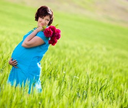 Happy pregnant woman with flowers outdoors, walking in field of green grass.