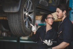 Young man and woman car mechanic wearing safety glasses with uniform and hand gloves standing under car in garage inspecting and doing maintenance work for car in garage