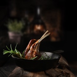 Traditional, roasted rack of lamb cutlets in a wrought iron skillet with rosemary herbs shot against a dark, rustic background with generous accommodation for copy space.