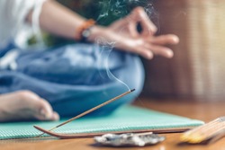 Woman meditating in lotus position on turquoise yoga mat on wooden floor. Focus on incense stick and smoke. Unrecognizable yoga practitioner in the background. Relax after Yoga training