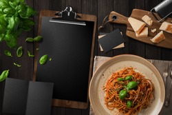 Blank restaurant menu template on dark wood table, with pasta, basil cheese, top view.