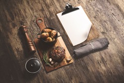 Steak on wooden serving board, with baked potatoes in a pot and sauce, with cliboard menu template, isometric view with copy space