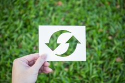 Hand holding cut paper with the logo of recycling over green grass. Recycling sign and symbol background banner concept, Earthday 