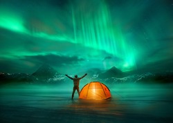 A man camping in wild northern mountains with an illuminated tent viewing a spectacular green northern lights aurora display. Photo composition.