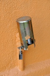 An outdoor water tap covered with steel cap and securely padlocked from theft with painted orange wall on background.