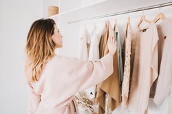 Cute young woman in bathrobe standing in front of hanger rack and trying to choose outfit dressing for work or walk. Selection of a wardrobe, stylist, shopping.