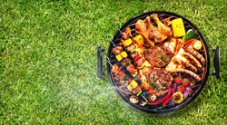 Top view of assorted delicious grilled meat with vegetables on barbecue grill with smoke and flames in green grass