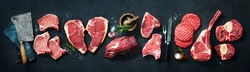 Assortment of raw cuts of meat, dry aged beef steaks and hamburger patties for grilling with seasoning and utensils on dark rustic board
