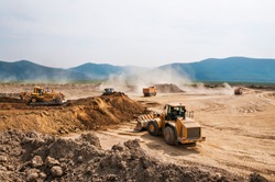  Earthworks on a summer day in a mountainous area. Wheel loaders, bulldozers and dump trucks are in operation. Dust from dump trucks that transport this soil
