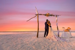 Bride and groom, newlyweds, honeymoon on the beach sunset sun under wedding arch with cake. Lovers or newlywed married young couple by the sea, wedding ceremony on a tropical beach, ocean background