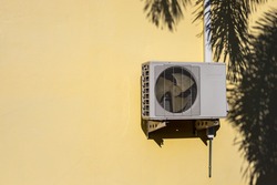 Close-up of the air conditioner compressor, which is installed on the yellow wall of the building near the shadow of the palm leaf silhouette.
