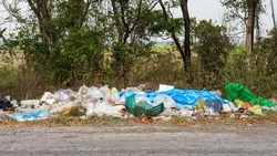 View of piles of plastic waste dumped on the side of a road near the trees and rice fields that are common in rural Thailand.