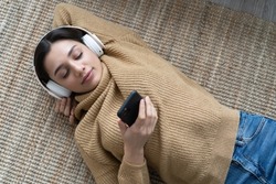 Woman in casual clothing with headphones lying down the floor in her living room and listening the music from her phone. Leisure time of single woman in isolation streaming songs from internet.