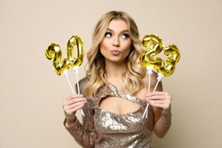 Charming young blonde girl with curly hair wearing sparkle dress holding balloons number 2023 in her hands on a beige background, new year holiday concept