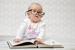 Funny portrait of a cute baby girl in glasses lying over a big book