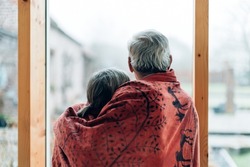 shot from behind of embraced elderly couple wrapped in a blanket looking out of a large glass wall