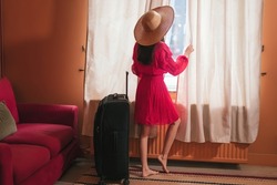 woman in hat leaving for vacation - lady dreaming of traveling looking outside the window - girl arrived in hotel room with luggage