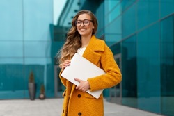 Business woman with laptop dressed yellow coat standing outdoors corporative building background Caucasian female business person eyeglasses on city street near office building Stylish businesswoman