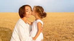 Daughter kisses her mom cheek standing wheat field. Little girl kiss mother Happy Mother's Day. Happy family relationship outdoor. Dressed white. Caucasian female woman 35 years and girl 5 years old