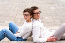 Portrait of two attractive students man and woman wearing stylish glasses sitting floor outdoor and smiling, head to head cheek to cheek, break relax between lesson, work, projects Eyeglasses concept