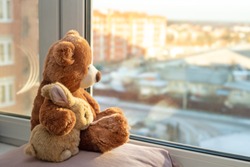 Best friends teddy bear and bunny toy sitting on window sill hugging each other and looking out of windows, sunlight, sunny day. Side view. Love, family and friendship concept. stay at home, safe.