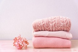 Stack of folded wool knitted sweaters or pullovers in pink pastel colors on table with flower hydrangea. Close up of warm cozy comfortable monochrome clothes for autumn winter season.