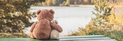 Stuffed toys brown teddy bear hugs bunny sitting on green bench on bank of tranquil lake on autumn day back view. Banner for web site.