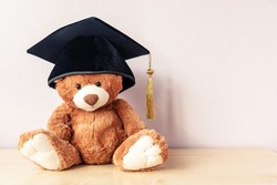 Adorable teddy bear with black academic cap sits on wooden table near light pink wall as space for text. Education for students