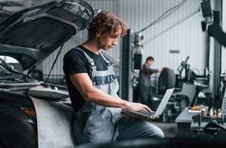 With laptop. Adult man in grey colored uniform works in the automobile salon.