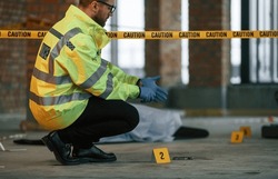 Wearing gloves. Male detective is collecting evidence in a crime scene near dead body on the construction site.